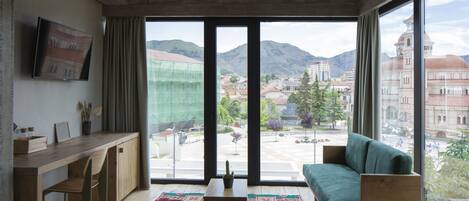 A modern living room with a wooden desk, a green sofa, and a large window offering a view of a cityscape and mountains.