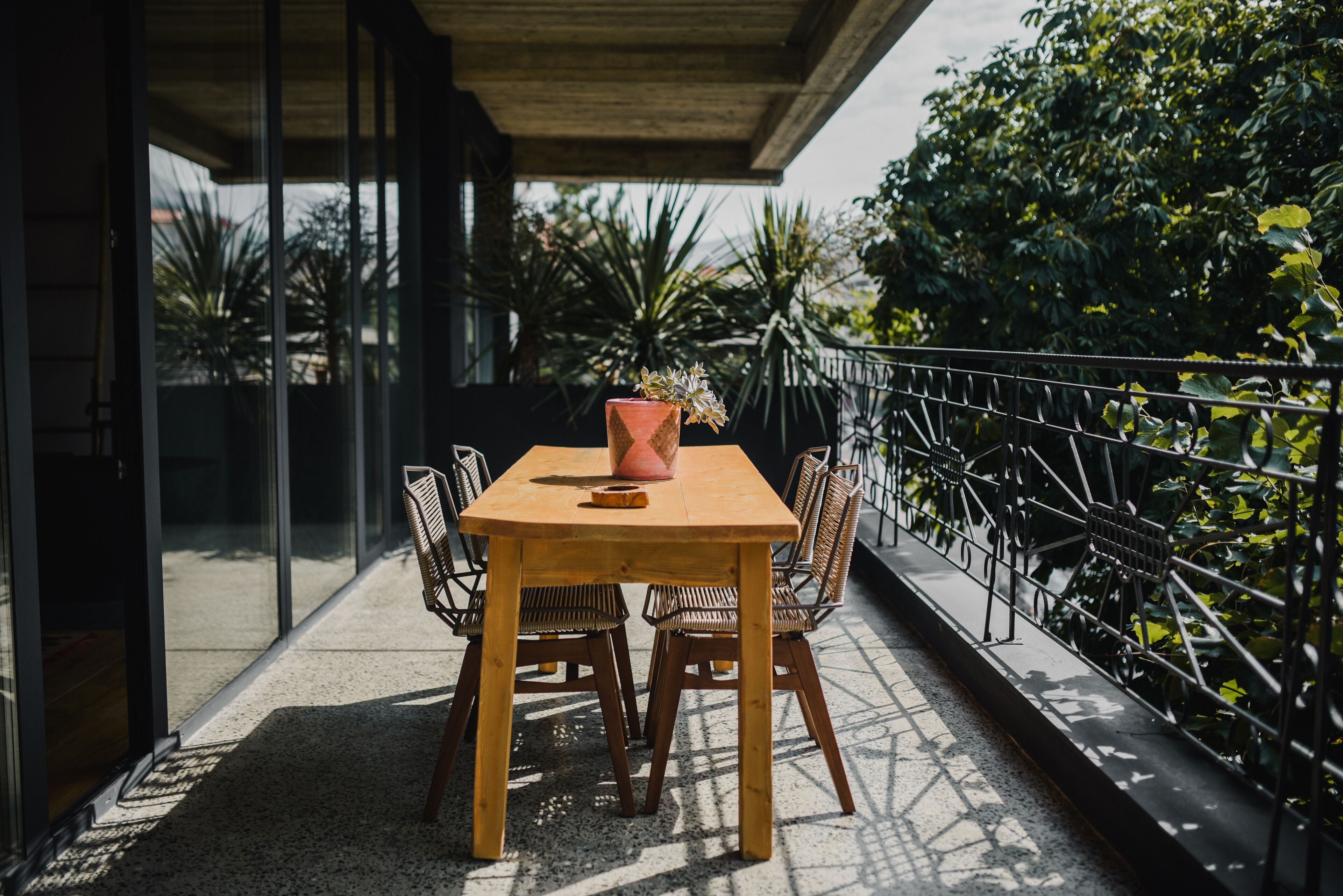 A wooden table with chairs on a balcony with a metal railing, surrounded by greenery.