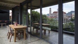 A modern outdoor dining area with a wooden table and chairs, large glass windows, and a view of a historic building with a tower. - In Gallery Spa & Hotel (Korçë)