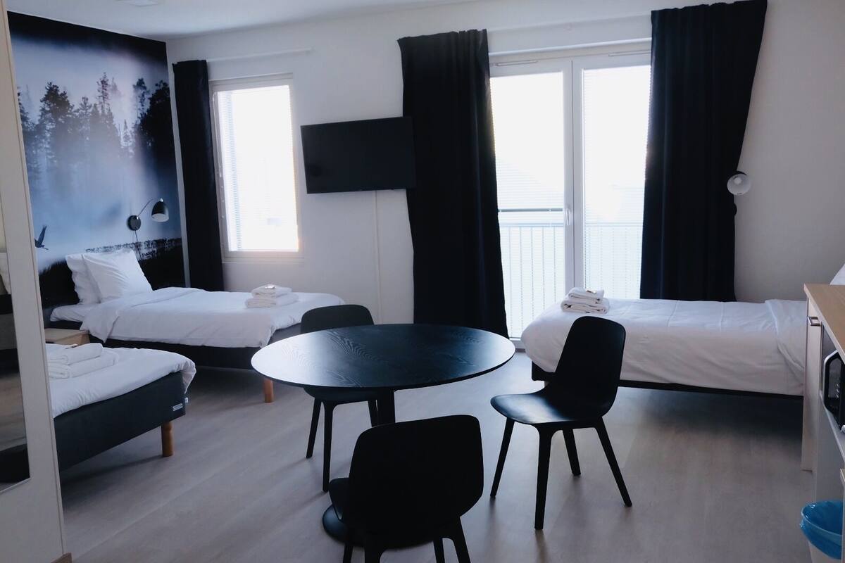 A hotel room with two beds, a round table, and chairs. There is a TV mounted on the wall, and a window with curtains.