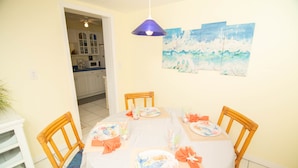 Een eetruimte met een ronde tafel voor vier personen, omringd door houten stoelen. Daarboven hangt een plafondventilator en aan de muur hangt een schilderij van een zeegezicht.