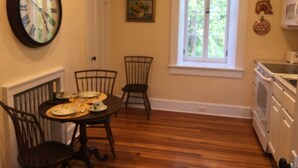 A dining area with a wooden table set for four, two chairs, a clock, a window with a floral curtain, and a kitchen area with appliances. - The Castle - c. 1780 (Lexington)