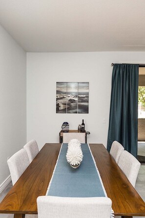 Un comedor con una mesa de madera, camino azul y sillas blancas. Se observa una obra de arte colgada en la pared y una mesita con botellas.