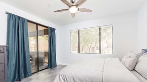 Un dormitorio con cama, ventilador de techo, ventana con persianas y puerta corredera de vidrio que da a un espacio al aire libre.
