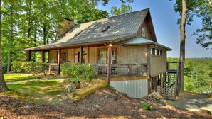 A rustic cabin with a covered porch, a stone chimney, and a view of the forest.