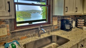 A kitchen with a stainless steel sink, a coffee maker, and a window with a view of a lake and mountains.