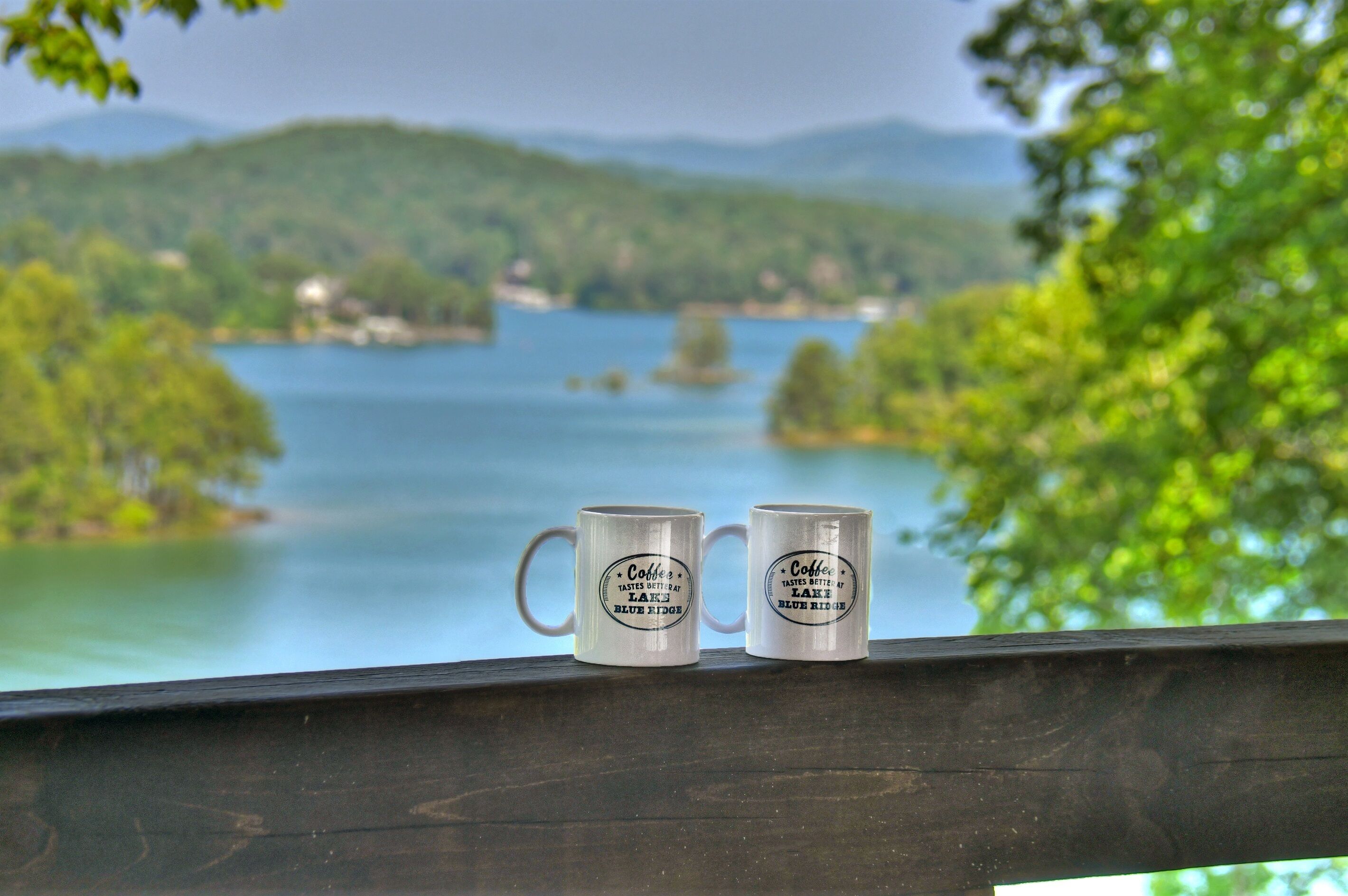 Dos tazas de café sobre una baranda de madera, con una vista panorámica de un lago y colinas al fondo.