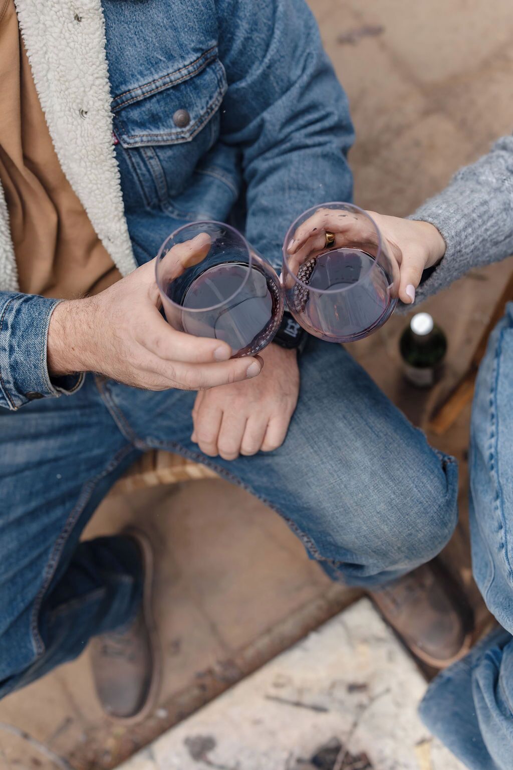 Dos personas sosteniendo copas de vino tinto, sentadas sobre una superficie de madera con una botella al fondo.