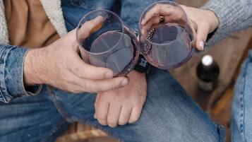 Dos personas sosteniendo copas de vino tinto, sentadas sobre una superficie de madera con una botella al fondo.