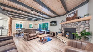 A spacious living room with a stone fireplace, wooden ceiling beams, and a large window overlooking a deck. - The Sparrow’s Nest ~ Cozy Charm + Fire Pit Fun (Shaver Lake)