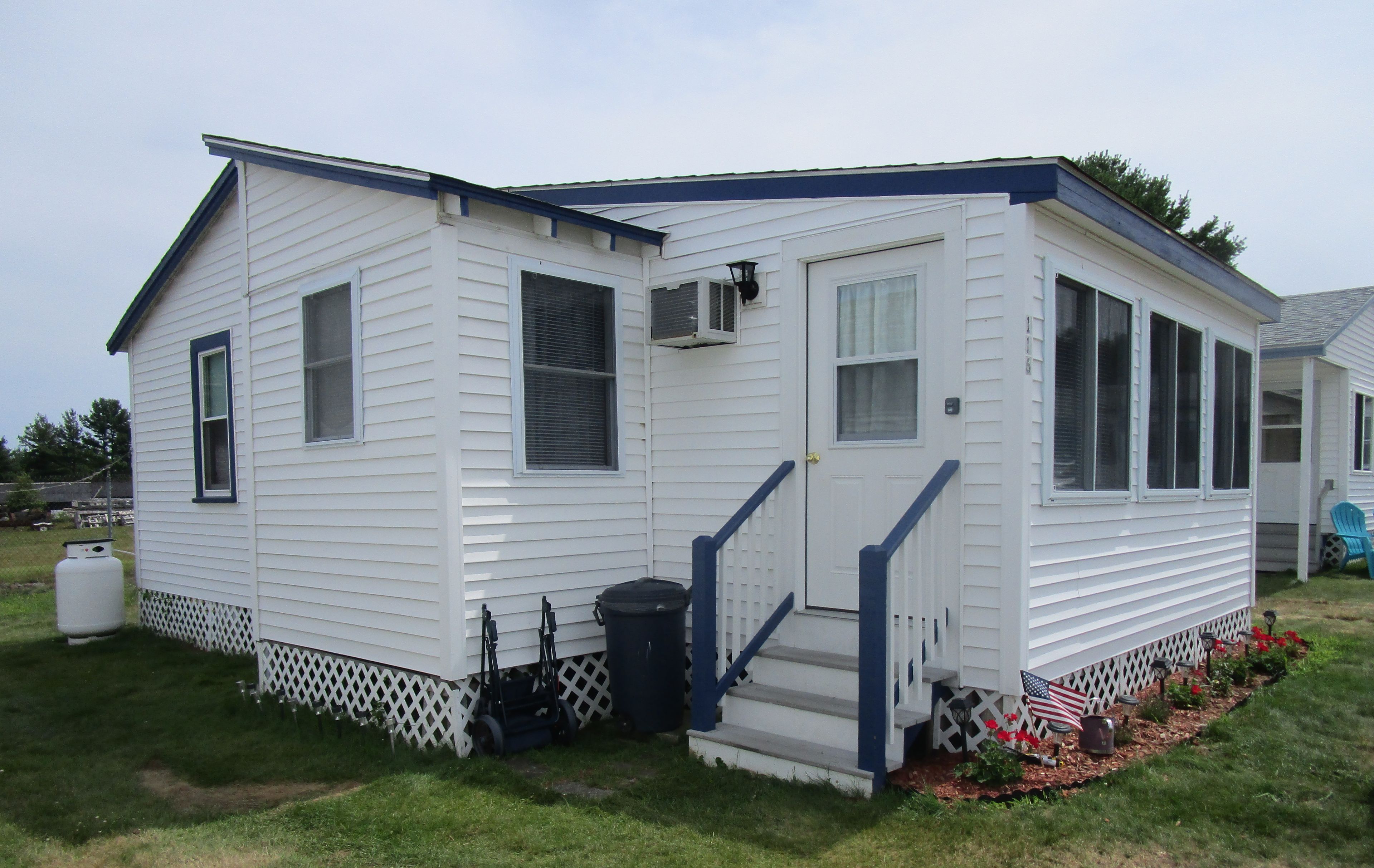 A small, single-story mobile home with a front porch, a small deck, and a front yard with a garden.