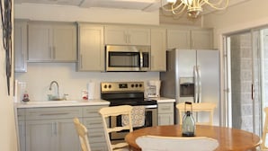 Una cocina con gabinetes gris claro, un microondas de acero inoxidable, una estufa y un refrigerador. Hay una mesa de comedor de madera con sillas y una lámpara de araña colgando.