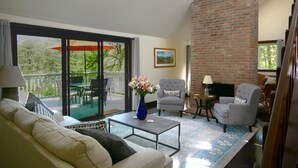 Ein Wohnzimmer mit Sofa, Sesseln, einem Couchtisch und einem Kamin. Es gibt einen Balkon mit Gartenmöbeln und ein großes Fenster.