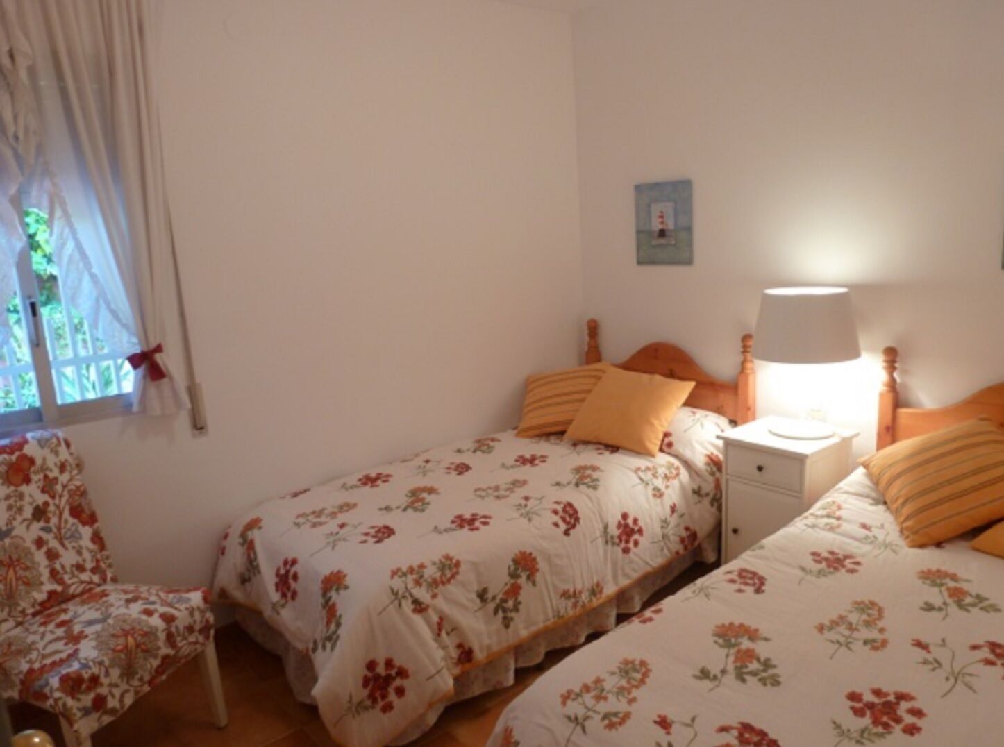 A bedroom with two single beds, a chair, a bedside table with a lamp, and a window with curtains.
