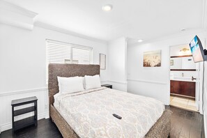 A hotel room with a bed, a nightstand, a small table, a window with blinds, a painting on the wall, and a bathroom visible through an open door.