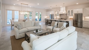 A spacious living room with a white sofa set, a coffee table, and a dining area with chairs. The kitchen features stainless steel appliances and pendant lighting.