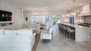 A spacious living area with a white sofa set, a dining area with chairs, a kitchen with modern appliances, and a large TV mounted on the wall.