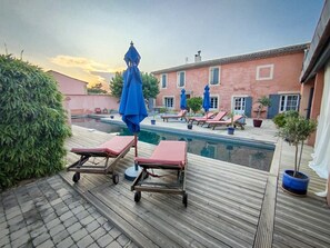 Un área junto a la piscina con sillones de descanso y sombrillas, una terraza de madera y un edificio con ventanas al fondo.