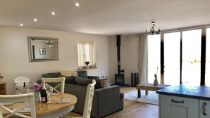 A cozy living room with a sofa, a dining table with chairs, a kitchen area with cabinets, and a large window with curtains. - Countryside Home with jacuzzi hot tub, open plan living and wood burner. (Ruthin)