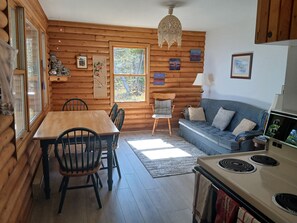 Ein rustikaler Innenraum einer Blockhütte mit Esstisch, Stühlen, einer Couch, einem Kamin und einem Fenster mit Blick auf Bäume.