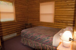 Ein Schlafzimmer in einer Blockhütte mit einem Bett, einem Nachttisch mit Lampe und zwei Fenstern mit Jalousien.