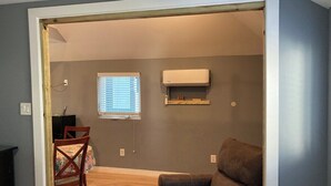 An open-plan living space with a brown leather sofa, a wooden dining table with a floral tablecloth, a window with blinds, and a wall-mounted air conditioning unit. - The GREEN Tiny House (Houston)