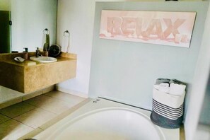 Une salle de bain avec un meuble-lavabo beige, un lavabo blanc, un miroir, un porte-serviettes et un cadre mural sur lequel est inscrit le mot « RELAX ».