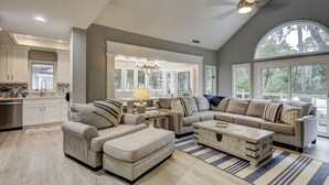 A spacious living room with a high ceiling, large windows, a kitchen area, and a dining setup.