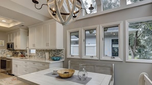 A modern kitchen with white cabinetry, a large island, and a view of the outdoors through multiple windows.