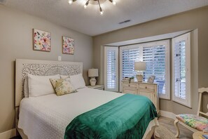 Une chambre à coucher avec un grand lit, une table de chevet, une commode et une fenêtre avec des stores.