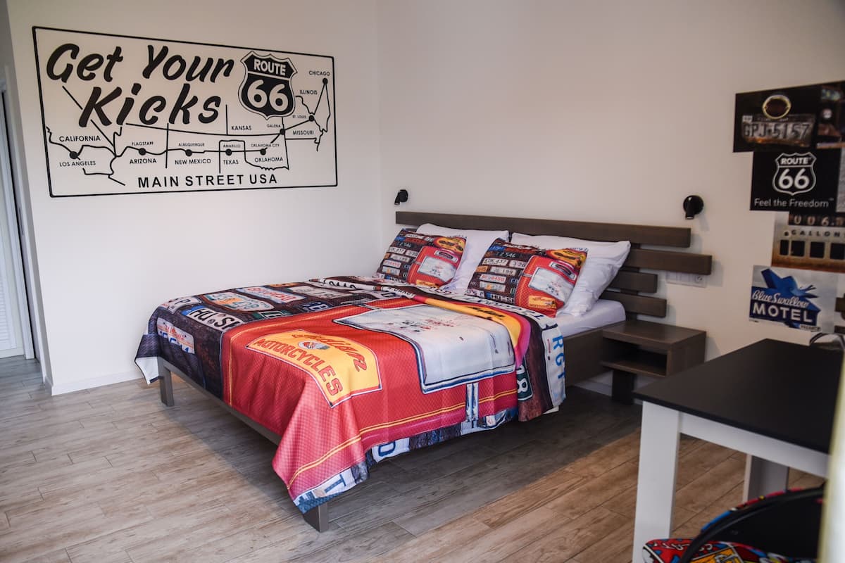 A bedroom with a Route 66 themed bedspread, a map on the wall, and a small table with a suitcase.