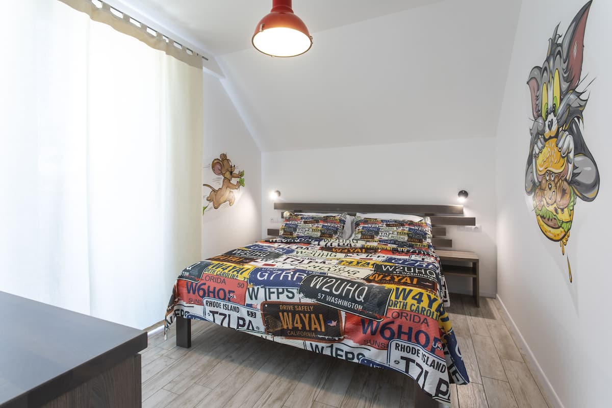 A bedroom with a bed covered in a blanket with various license plates, a wall mural of a cartoon character, and a staircase leading to another room.