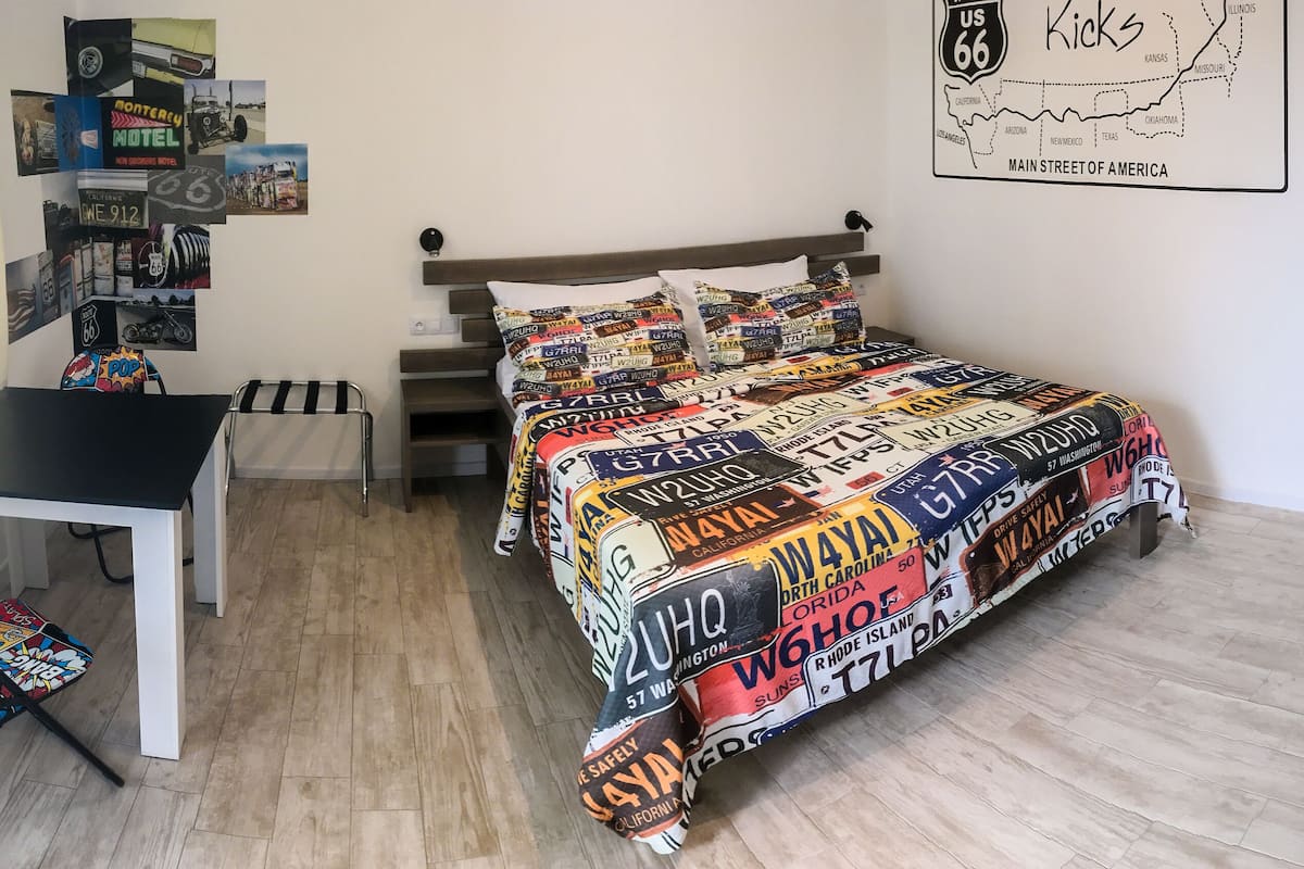 A bedroom with a bed covered in a California license plate print, a small table, a chair, and a wall with a map and various posters.