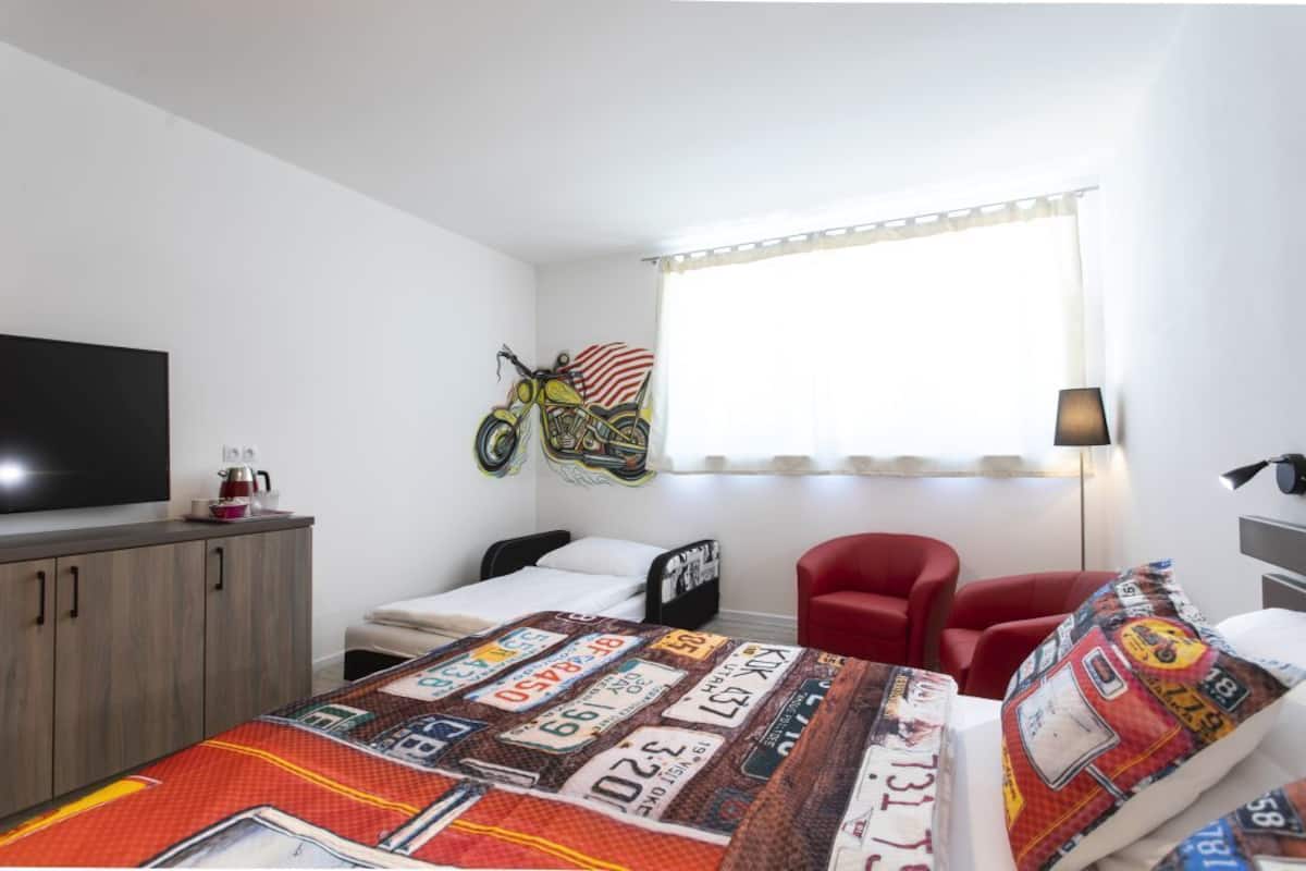A compact hotel room with a bed, a red chair, a television, and a motorcycle decoration on the wall.