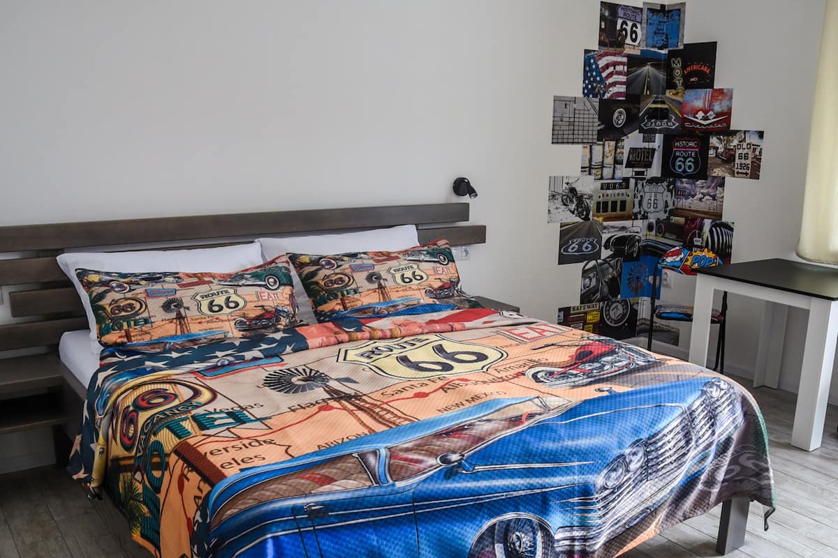 A bedroom with a bed covered in a Route 66 themed blanket, a wall decorated with Route 66 memorabilia, and a nightstand with a lamp.