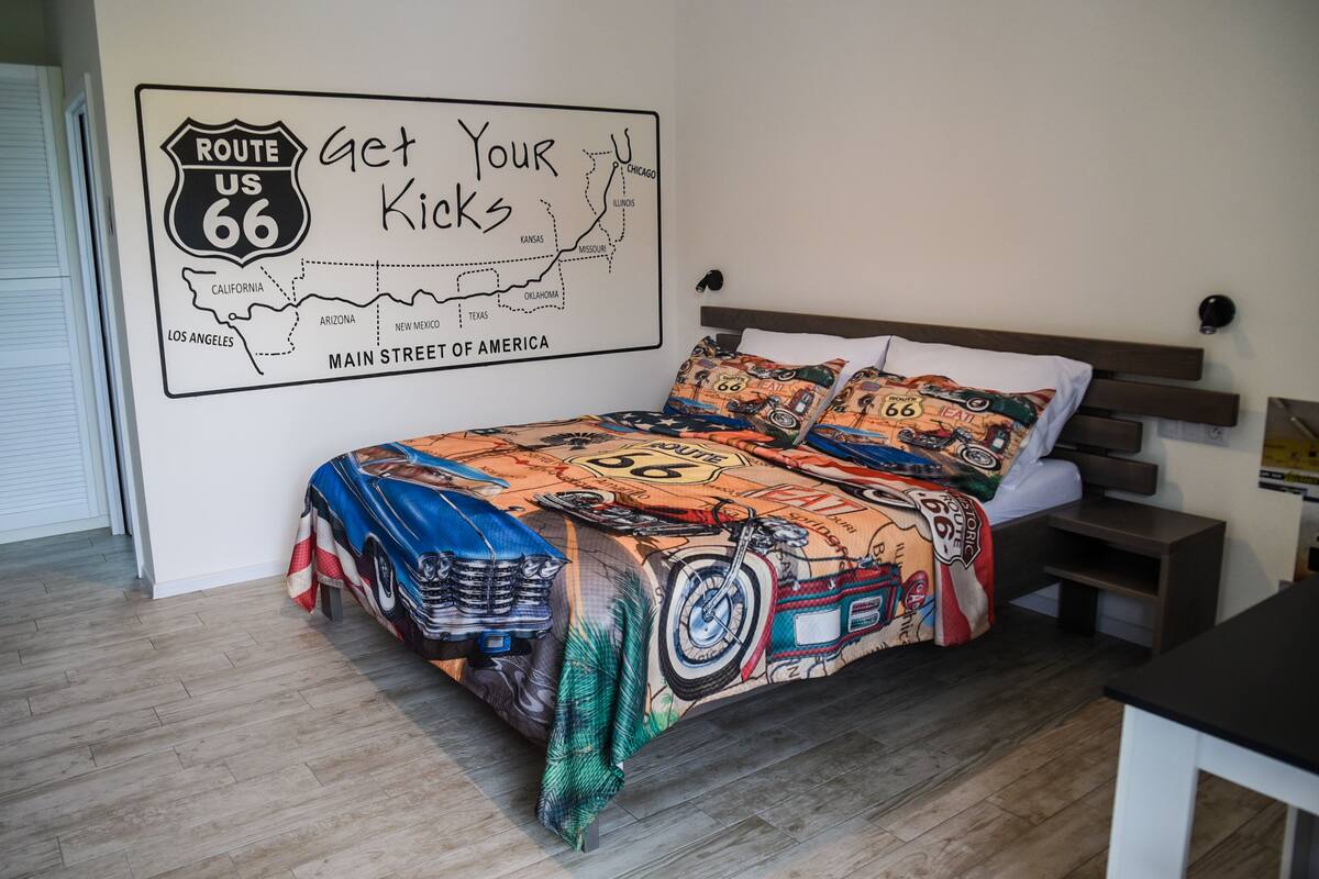 A bedroom with a bed featuring a Route 66-themed bedspread, a wall map of Route 66, and a wooden headboard.