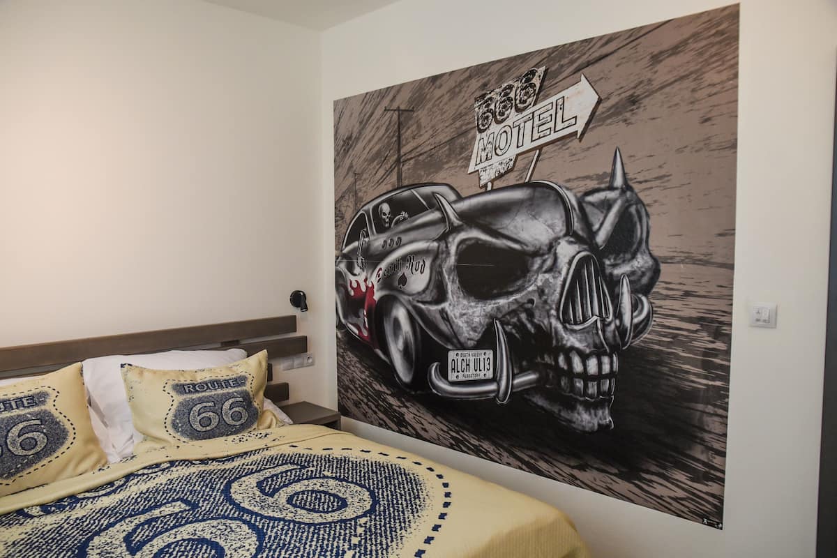 A bedroom with a bed, pillows, and a large wall mural featuring a skull and a car.