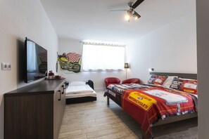 A bedroom with a bed, a desk, a television, a motorcycle-themed bedspread, and a window with curtains. - Motel Eldorado (Mikulov)
