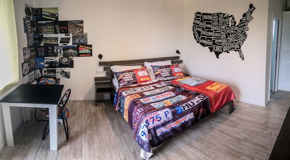 A bedroom with a bed covered in a blanket featuring various U.S. state license plates, a wall map of the U.S., and a collection of vintage travel posters on the wall. - Motel Eldorado (Mikulov)