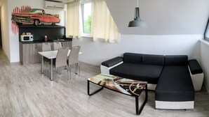 A modern living space with a black sectional sofa, a dining table with chairs, a kitchen area with a microwave, and a large window with curtains. - Motel Eldorado (Mikulov)