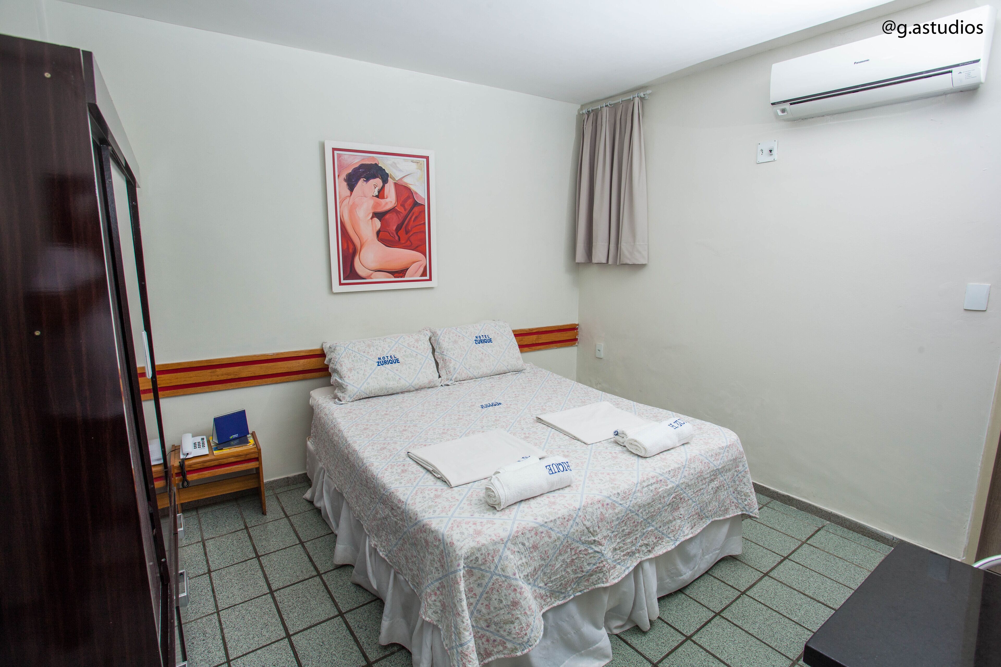 Una habitación de hotel con cama, mesita de noche, unidad de aire acondicionado y un cuadro en la pared.