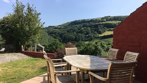 Eine Terrasse im Freien mit einem runden Holztisch und vier Stühlen, mit Blick auf eine üppige, grüne Landschaft mit Hügeln.