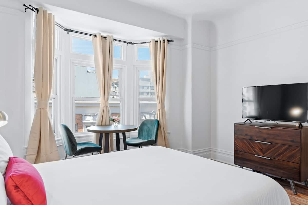 Une chambre d’hôtel comprenant un lit, une télévision, une commode, une fenêtre avec des rideaux, une petite table et deux chaises.