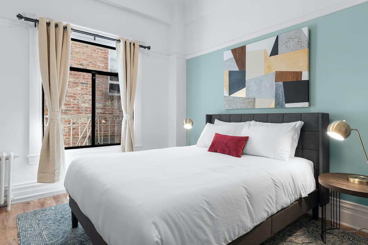 Une chambre moderne avec un grand lit, une vue sur un immeuble en briques par la fenêtre et un tableau abstrait aux motifs géométriques accroché au mur.