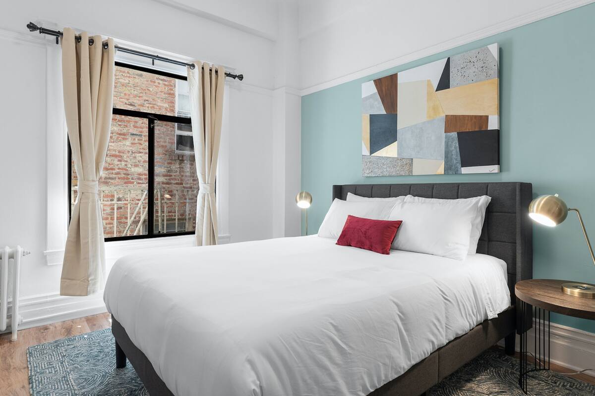 Une chambre moderne avec un grand lit, une vue sur un bâtiment en briques et une œuvre d’art géométrique accrochée au mur.