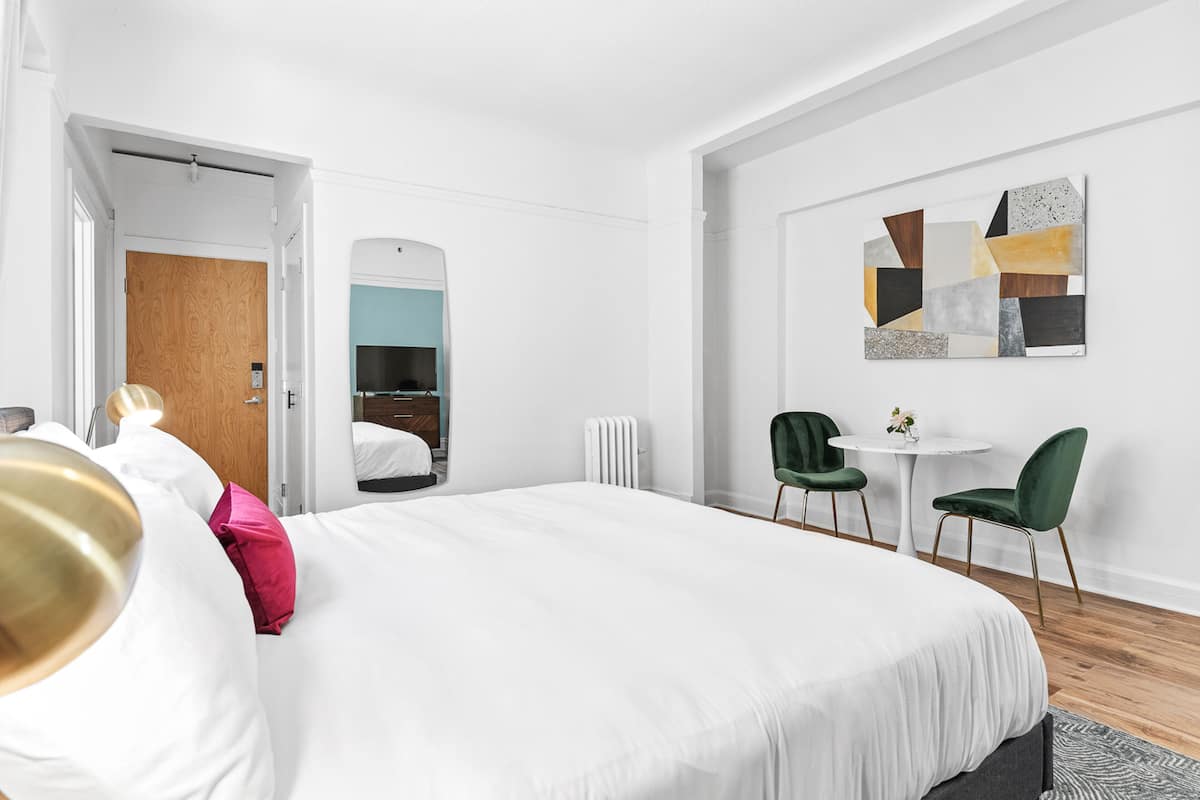 Une chambre d’hôtel moderne avec un grand lit, une petite table avec des chaises, un miroir et une œuvre d’art murale.