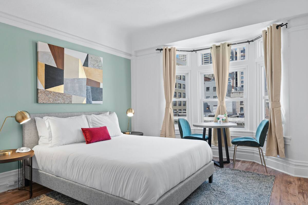 Une chambre d’hôtel avec un lit, un bureau, une chaise, une fenêtre avec des rideaux et une vue sur la ville.