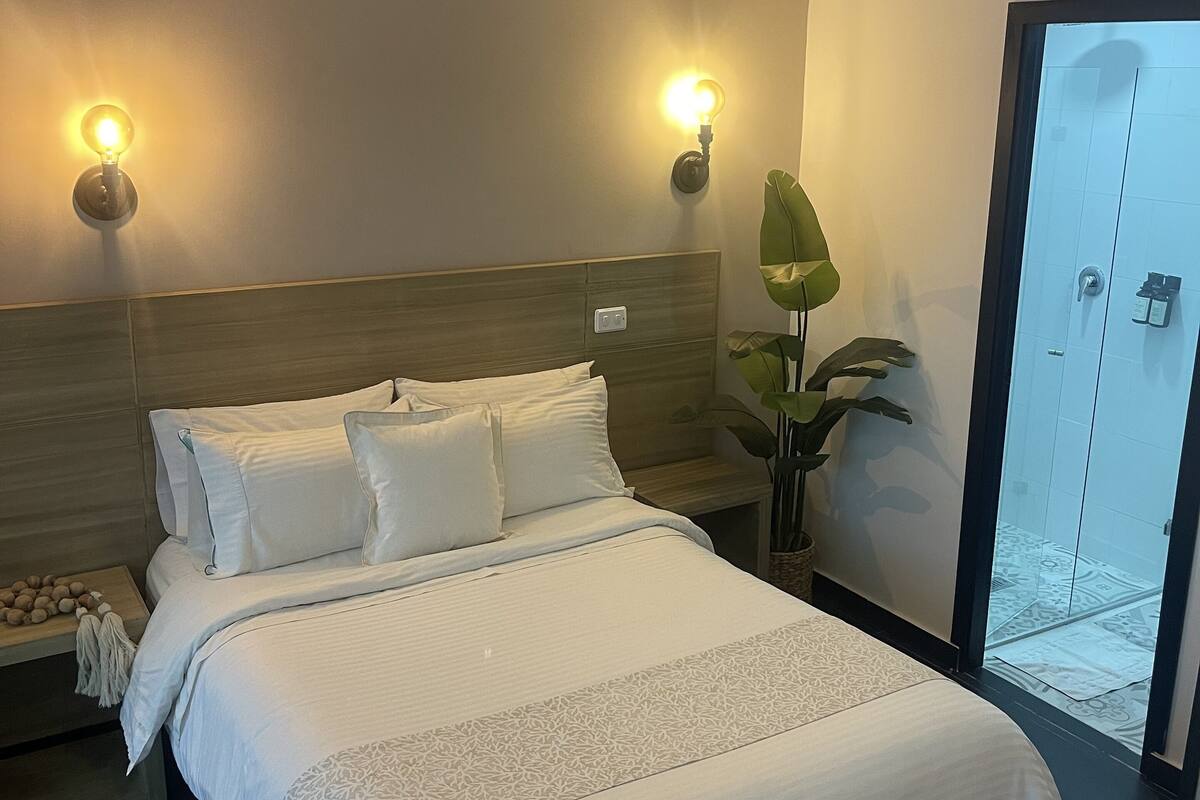 Ein Hotelzimmer mit einem großen Bett, zwei Wandlampen, einer grünen Pflanze und einer Schiebetür zu einem Badezimmer.