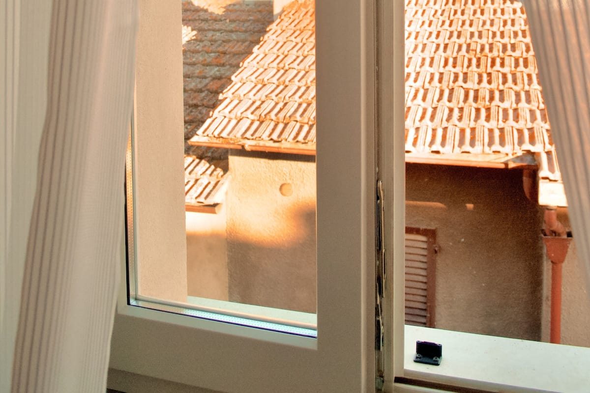 A window with curtains, revealing a view of a building with a tiled roof and a chimney.