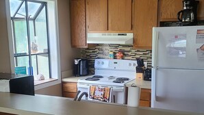 A kitchen with wooden cabinets, a white refrigerator, a stove, and a coffee maker. There are three wooden stools at the counter. - Cozy 2 bed 2 bath in Arcadian Dunes (Myrtle Beach)